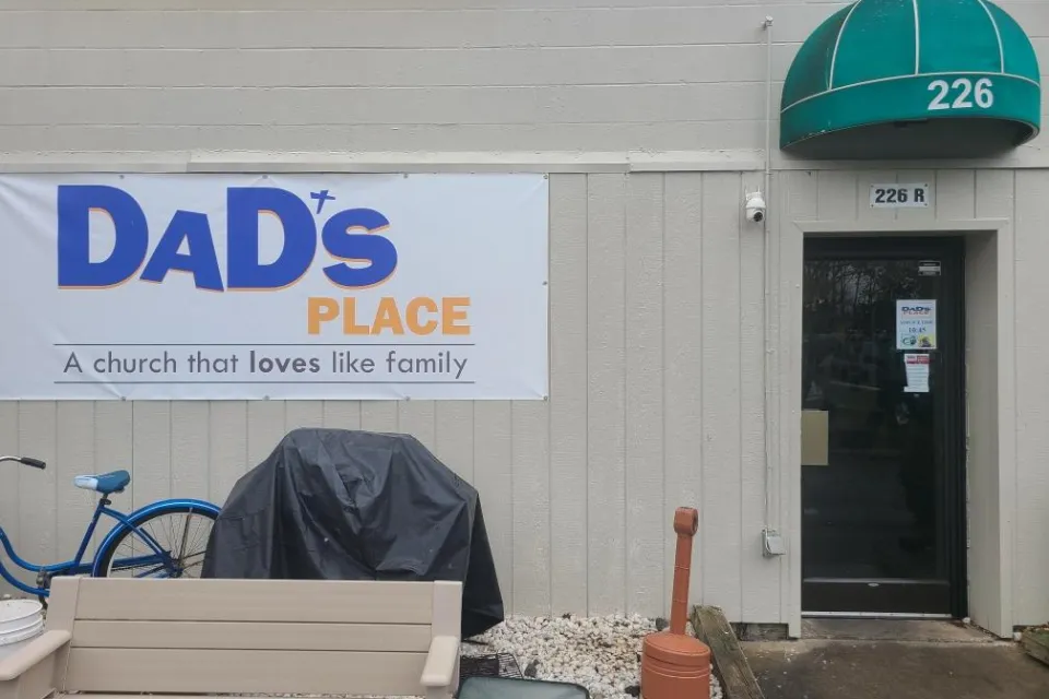 White sign on side of tan building with metal siding and green overhang, reading "Dad's Place, a church that loves like family."